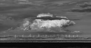 Black and white image of a landscape with numerous wind turbines spread across an open field. Large, dramatic clouds dominate the sky, creating a striking backdrop. The scene conveys a sense of vastness and renewable energy potential.