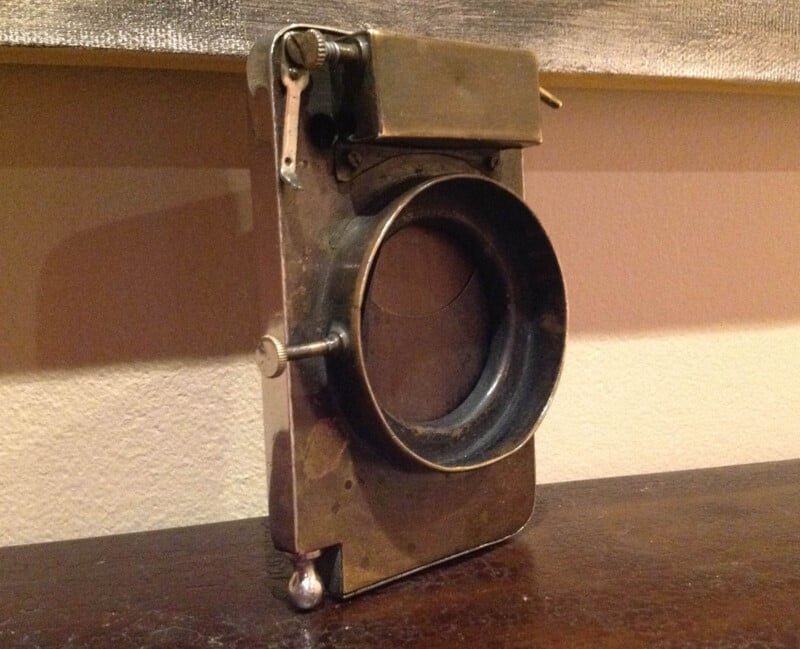 A vintage brass and metal camera lens on a wooden surface, with intricate mechanical details and a rectangular, box-like structure. The lens has a large circular opening, and is slightly tarnished, indicating age and use.