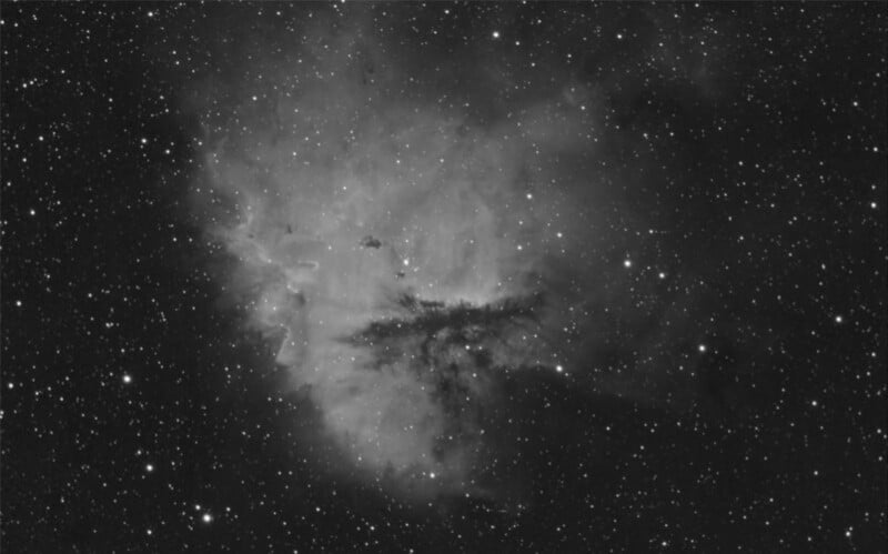 A black and white image of a dense star field featuring a bright, cloud-like nebula. The nebula has intricate patterns and varying shades of grey against the black background, creating a vivid contrast with the surrounding stars.