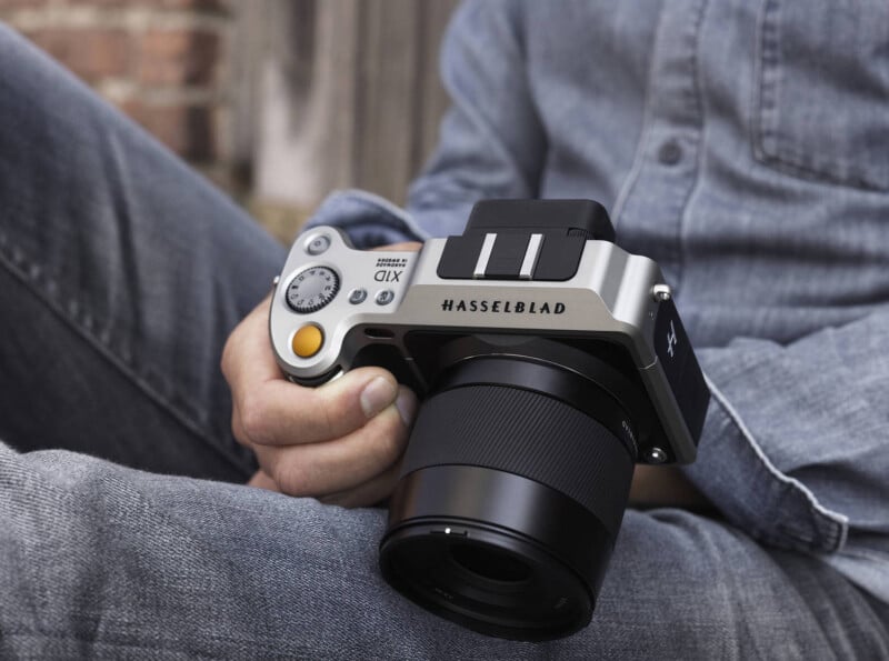 Person holding a Hasselblad X1D camera with one hand, dressed in a light denim shirt and jeans. The background is blurred, featuring some bricks and wood elements.