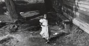 A young girl walks in a yard near a wooden building, holding a dress. The ground is uneven with grass and debris scattered. Shadows from trees and structures create a contrasting backdrop.