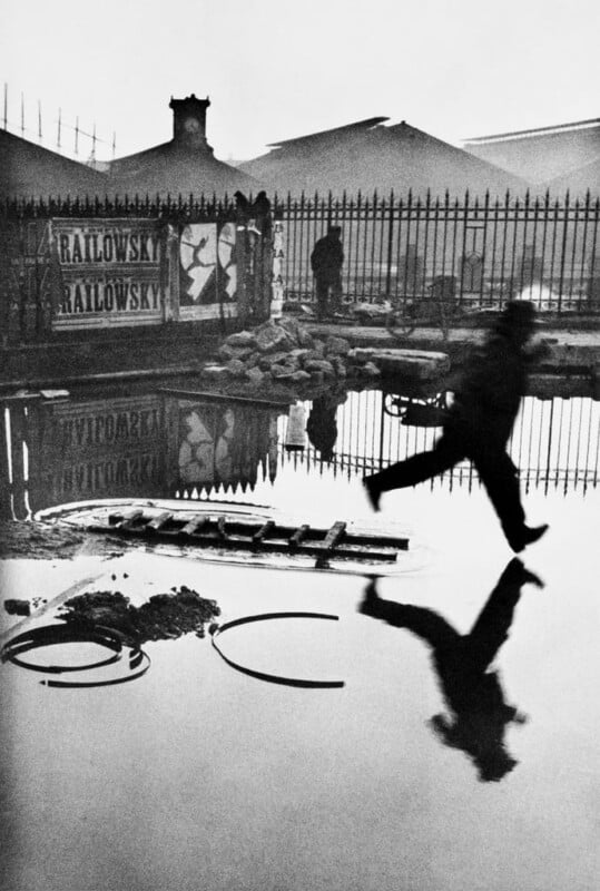 A blurred silhouette of a person leaps over a large puddle, casting a reflection in the water. A fence and a building with posters are in the background. The scene is in black and white, creating a dramatic and timeless atmosphere.