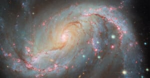 A vivid spiral galaxy with glowing arms extends outward, showcasing bright stars and clusters of pinkish-red nebulas against a dark space backdrop. The galaxy's spiral structure is prominent and intricate.