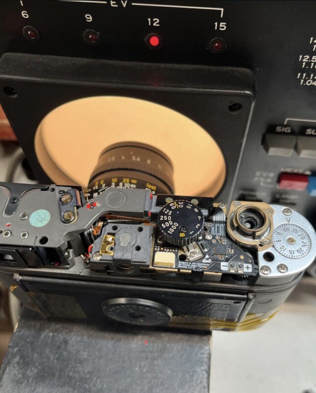 A close-up of a disassembled vintage camera on a table, displaying its mechanical and electronic components. The camera is positioned against a circular backlight from a piece of equipment with dials and buttons visible in the background.