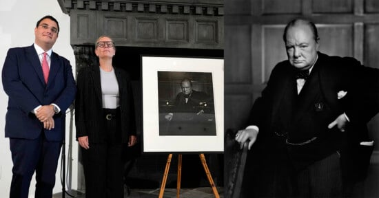 Two individuals stand beside an easel displaying a black-and-white photograph of a man in formal attire. To the right, the same man is shown posing with one hand on his hip. The background features a dark paneled wall.