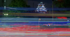 A night cityscape featuring a brightly lit traditional Japanese castle in the background. Colorful light trails from passing vehicles create dynamic streaks in the foreground, capturing the motion and energy of the scene.