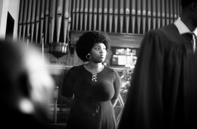 A black and white photo of a woman standing in a church. She is wearing a dark dress and hoop earrings, with an organ in the background. The setting is solemn, and other blurred figures can be seen nearby.