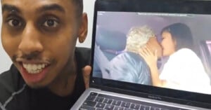 A person smiling and holding a laptop displaying a video. In the video, two people are seen kissing; one has blonde hair and the other has long brown hair.