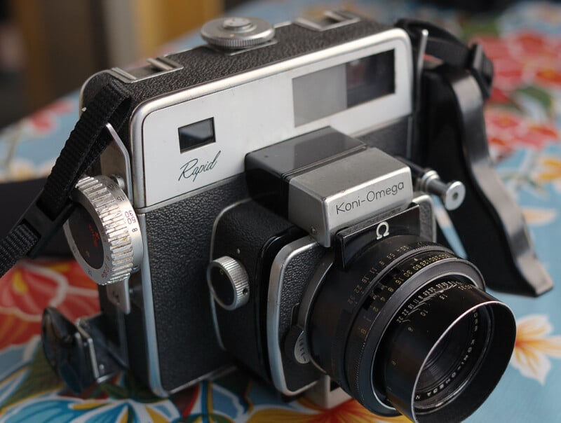 A vintage Koni-Omega Rapid camera with a black body and metallic details, featuring a large lens and multiple dials, rests on a colorful floral surface.