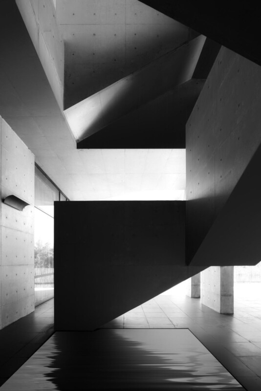 Black and white photograph of a modern interior with concrete walls and angular architectural elements. Large windows let in natural light, casting geometric shadows on the surfaces. The setting feels minimalistic and open.