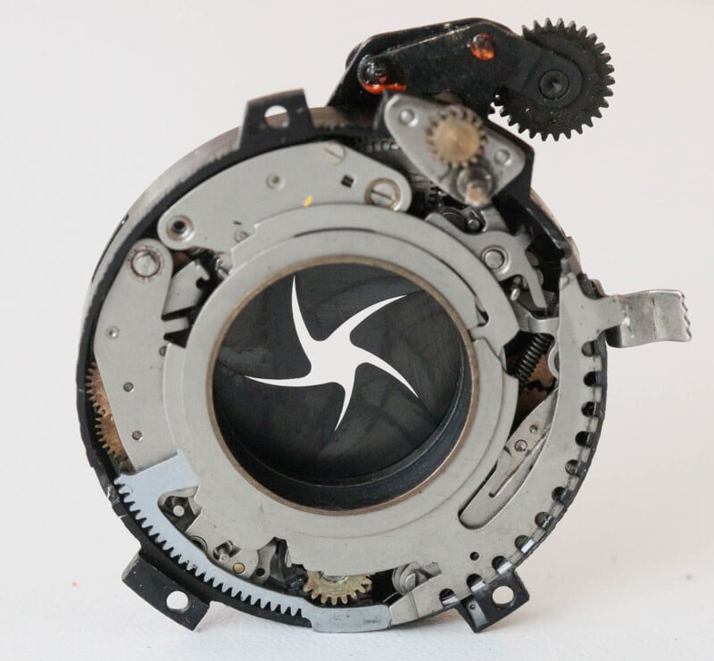 Close-up of a mechanical camera shutter assembly showing various gears, springs, and metal components. The central aperture features a star-shaped opening, while the surrounding parts exhibit intricate engineering details in silver, black, and bronze tones.