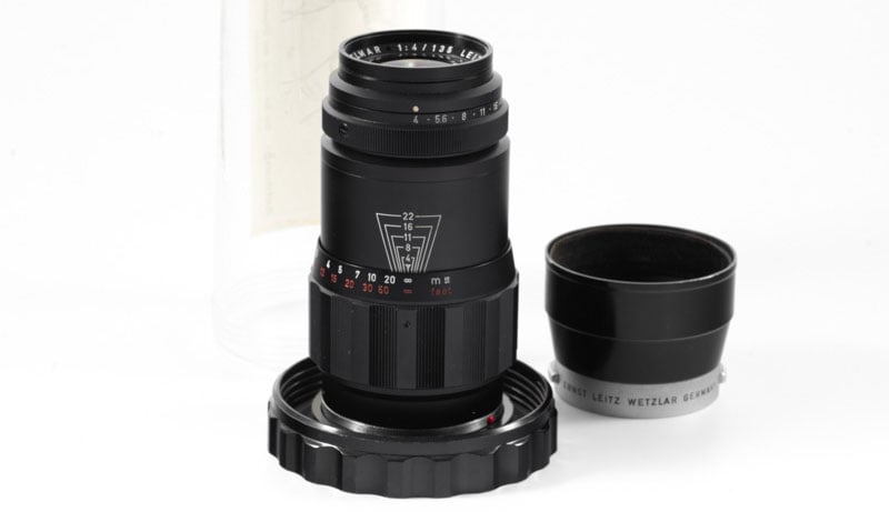 A black camera lens with a wide focus ring is placed upright on a white surface. The lens has engraved numbers for focus distances. Next to it is a detached lens hood, and behind it is a clear cylindrical container.