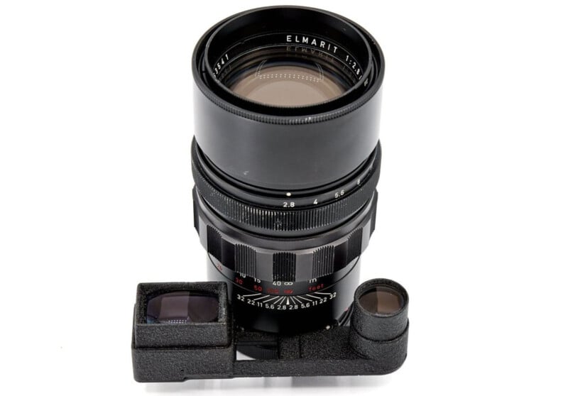 A vintage camera lens with a black body, featuring aperture and focus rings marked with numbers. It has a viewfinder attachment at its base, consisting of a rectangular and a circular element. The lens is labeled "Elmarit.