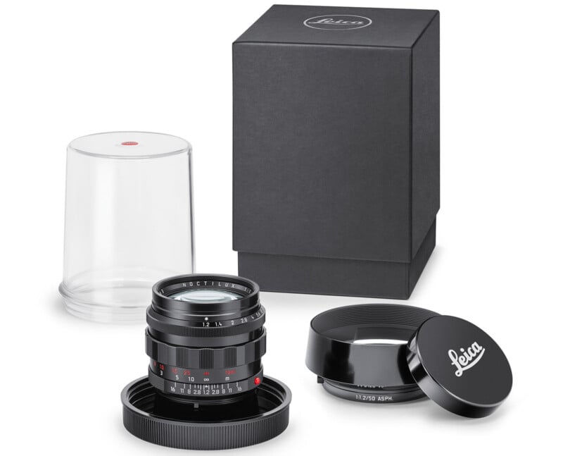 A Leica camera lens set including a lens with caps, a lens hood, a transparent plastic container, and a black rectangular box is arranged on a white background.