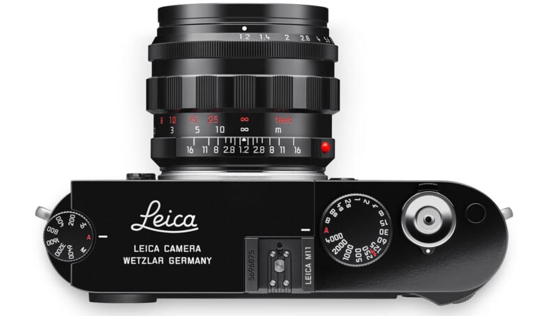 Top view of a black Leica camera with prominent dials and buttons, featuring the brand name and location engraved. The lens is attached, showing focusing and aperture scales in white and red lettering.