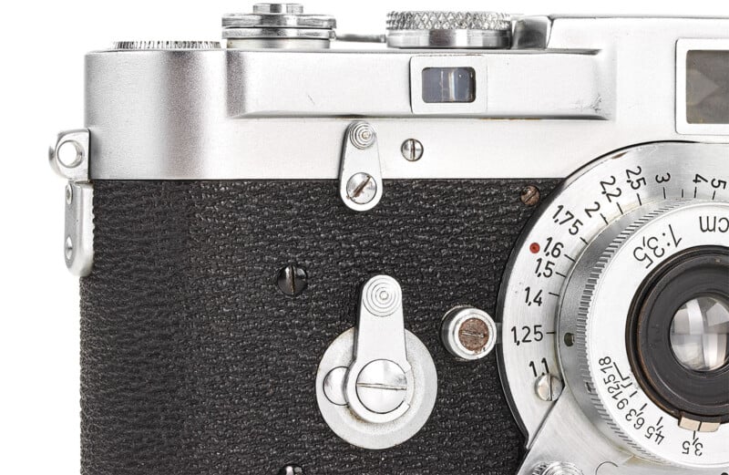 Close-up of a vintage silver and black camera, showing details of the lens, control dials, and textured grip. The lens has engraved numbers and markings, indicating focus settings. The camera features a classic design with mechanical components.