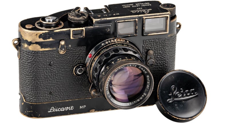 A vintage Leica MP film camera with a worn black and silver finish, featuring a lens with a cap placed beside it. The text on the cap and camera body reads "Leica." The camera has a textured body and visible dials and controls.