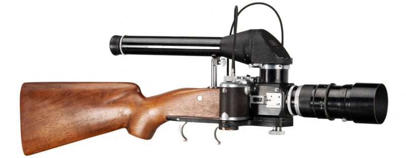 A vintage camera styled like a rifle, featuring a wooden stock and a long lens. The main body is black with metallic components, and a wire extends from the top. The overall design mimics a firearm.