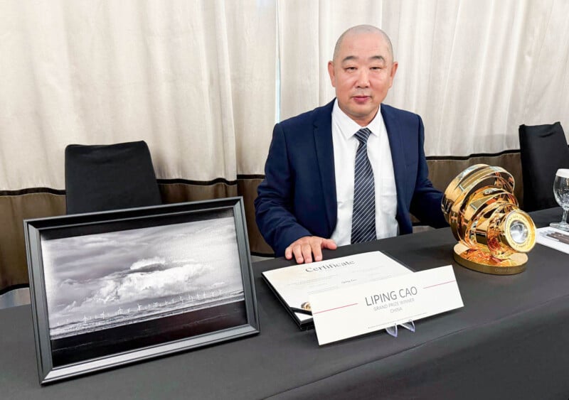 A man in a suit sits at a table with a gold trophy, a framed black and white photo, and a certificate labeled "Liping Cao.