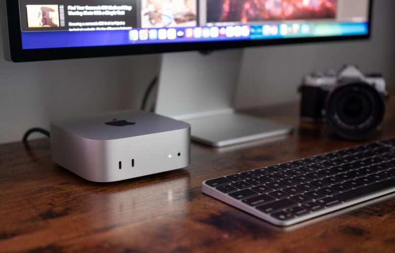 A sleek desktop setup featuring a modern computer monitor and keyboard on a wooden desk. Next to the monitor is a compact, silver computer with the Apple logo. A camera is positioned near the monitor, creating a tech-focused workspace scene.