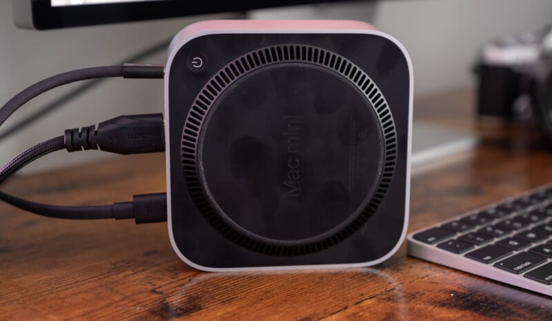 A Mac Mini placed upside down on a wooden desk, showing its circular base with the words "Mac mini." Cables are connected to the back, alongside a keyboard and part of a monitor in the background.