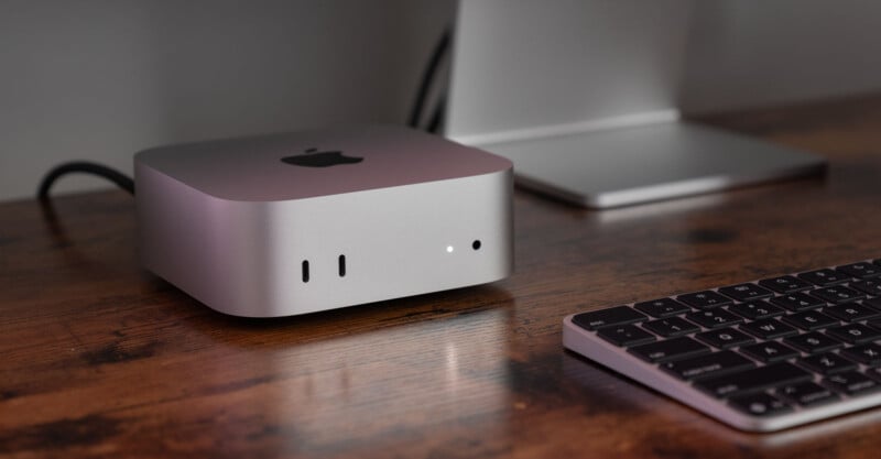 A compact desktop computer with an Apple logo sits on a wooden desk next to a keyboard. A monitor is partially visible in the background.