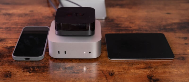 A smartphone, a compact square computer with a black device on top, and a large black trackpad are placed side by side on a wooden surface.