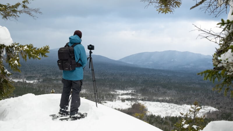 A person in a blue jacket, wearing snowshoes and a backpack, stands on a snowy cliff setting up a camera on a tripod. They overlook a vast, snow-covered forest with mountains in the distance under a cloudy sky.