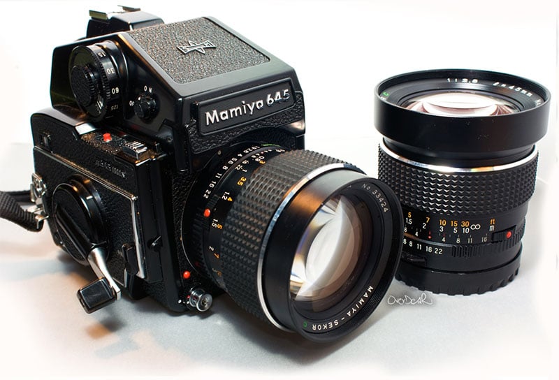 A Mamiya 645 camera with a large attached lens is positioned on a flat surface. Another separate lens sits beside the camera. Both lenses have detailed markings and focus rings, showcasing a classic, professional design.