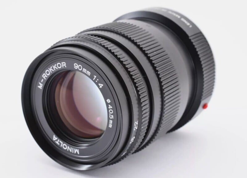 A Minolta M-Rokkor 90mm f/4 camera lens placed on a white background, showing its focusing rings, aperture markings, and front glass element.