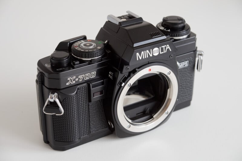 A Minolta X-700 camera without a lens is shown on a light background. The camera body features various control dials and buttons, with detailing in black and a metallic finish.