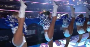 Cheerleaders in matching blue and white outfits perform high kicks in a stadium, holding blue and white pom-poms, with bright overhead lights and a large crowd in the background.