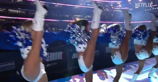 Cheerleaders in matching blue and white outfits perform high kicks in a stadium, holding blue and white pom-poms, with bright overhead lights and a large crowd in the background.