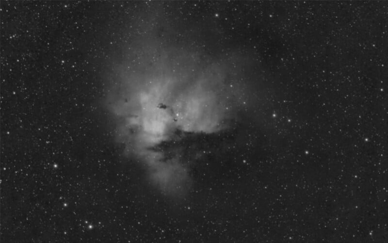 A black and white image of the Pacman Nebula, featuring a dense cluster of stars and cloud-like formations. The central region is brighter, surrounded by darker areas giving it a textured appearance against the star-filled background.