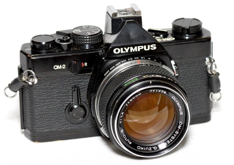 A black Olympus OM-2 film camera with a large lens is shown. The camera features dials and buttons on the top, and it has the name "Olympus" prominently displayed above the lens. Its overall design is vintage and classic.