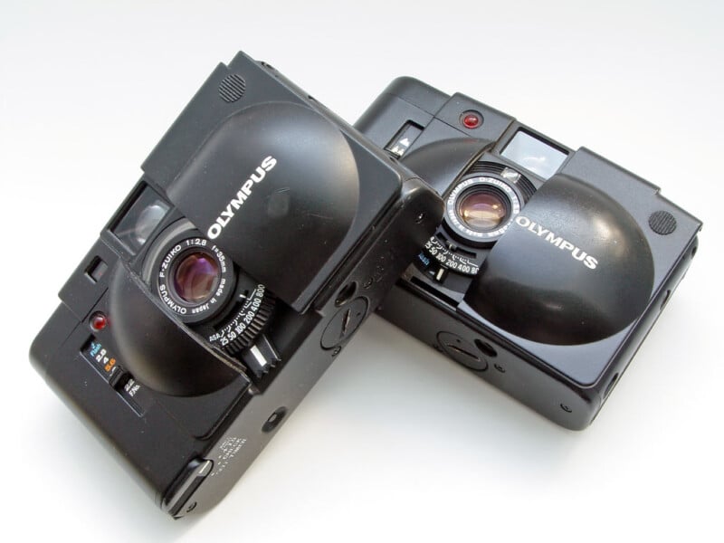 Two black Olympus XA compact film cameras displayed at an angle against a white background, showcasing their lenses and unique clamshell design.