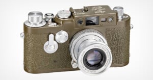 A vintage Leica camera with a textured olive green body and a silver lens. The top features various dials and buttons, and the lens is engraved with "Ernst Leitz GmbH Wetzlar.