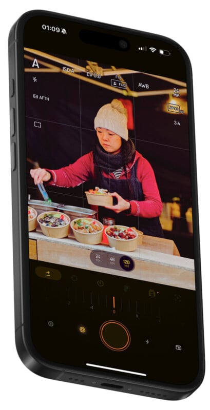 A smartphone screen displays a camera app focusing on a person in a beanie serving food into bowls at a counter. The image includes various camera settings and controls at the bottom.