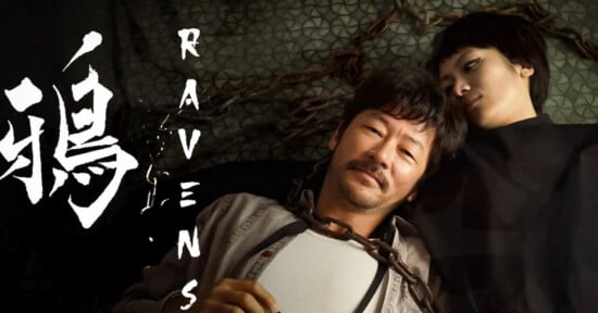 A man and a woman lie on the ground, with the man's head resting on the woman's shoulder. Chains are visible around the man's neck. The word "Ravens" and stylized characters are displayed on the left.