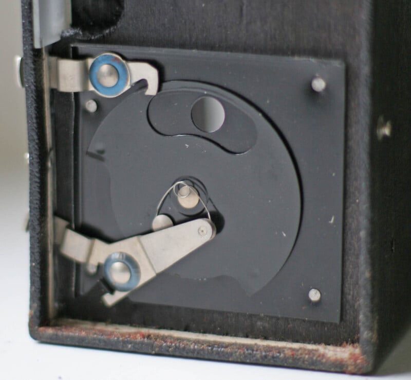 Close-up of a vintage camera’s internal mechanism, showing metal levers and a circular aperture inside a dark housing. The mechanical parts are silver against a black background.