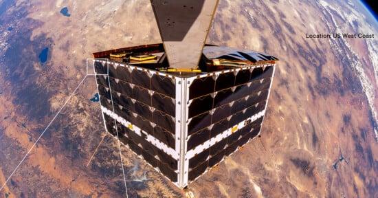 A satellite in orbit over the Earth with a backdrop of desert and mountains, indicated as the US West Coast. The satellite has a cube shape with solar panels, and part of its structural arm is visible.