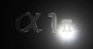 A chrome representation of the Greek letter "Alpha," the number "1," and the Roman numeral "II" is illuminated against a dark background with a bright light source on the right.