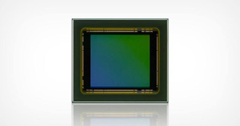 Square image sensor with a gradient from green to blue in the center, surrounded by a black border with gold contacts. The sensor is set against a light background, and its reflection is visible below.