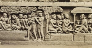 Ancient stone relief depicting a group of seated and standing figures in traditional attire. The scene includes trees and an architectural structure in the background, suggesting a historical or cultural narrative.