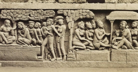 Ancient stone relief depicting a group of seated and standing figures in traditional attire. The scene includes trees and an architectural structure in the background, suggesting a historical or cultural narrative.