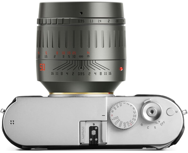 A top view of a camera with a large metallic lens attached. The focus ring is marked with distance and aperture measurements. The camera body has a silver finish with dials and buttons for settings adjustment.