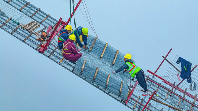 Construction workers in safety gear are working on a suspended section of a bridge against a clear blue sky. They are installing or inspecting metal grids, with cables and scaffolding visible around them.