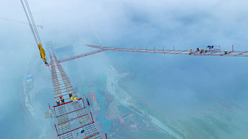 A high-angle view of a bridge under construction shrouded in mist. Workers in bright safety gear are on the narrow framework. Cranes are visible in the background, and construction materials are scattered below.