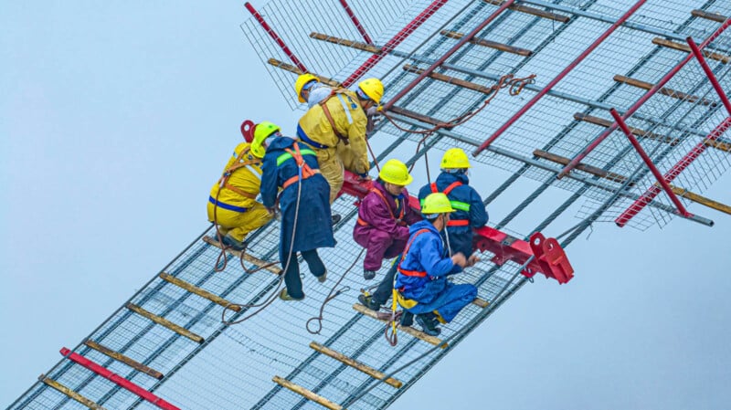 Construction workers in colorful safety gear work on a high, angled steel structure against a sky backdrop. They're using ropes and harnesses for safety, illustrating teamwork and precision in engineering.