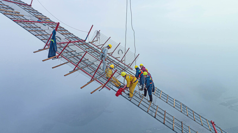 Workers in safety gear and yellow helmets position themselves on a suspended scaffolding high above the ground, against a cloudy sky. They appear to be engaged in a construction or maintenance task, using ropes and safety harnesses for support.